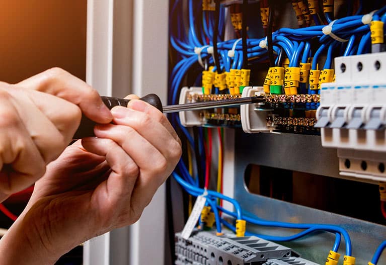 Électricien utilisant un tournevis sur un tableau électrique avec câblage bleu et disjoncteurs.